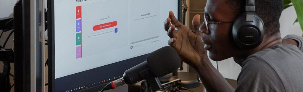 A man sits at a computer, speaking into a microphone and wearing headphones.